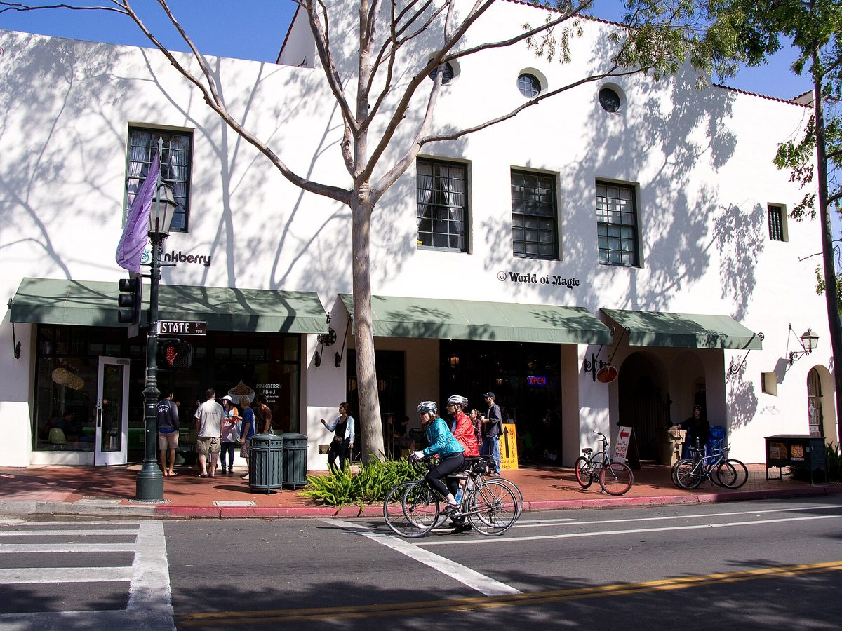 A white Spanish Colonial Revival building on State Street in downtown Santa Barbara, its shops under green awnings and cyclists at the crosswalk